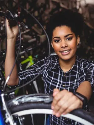 Eine junge Frau mit lockigem Haar und einem karierten Hemd lächelt, während sie den Lenker eines Fahrrads in einem Laden voller anderer Fahrräder hält.