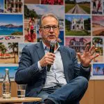 Ein Mann in einem blauen Anzug und mit Brille spricht in ein Mikrofon, während er auf der Bühne sitzt. Auf dem Tisch neben ihm stehen ein Glas und eine Flasche Wasser. Hinter ihm ist eine Collage mit bunten Reisefotos zu sehen.