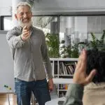 Ein lächelnder Mann mit Bart, der eine Brille und ein graues Hemd trägt, steht und zeigt auf eine Person, die ihre Hand in einem Klassenzimmer oder Büro mit Bücherregalen und Pflanzen im Hintergrund hebt.