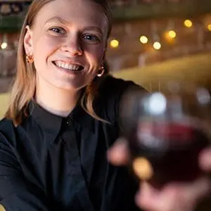Eine lächelnde Frau mit hellem Haar und schwarzem Hemd hält ein Glas Rotwein in die Kamera. Warme Lichterketten und eine Backsteinmauer sind im Hintergrund verschwommen.