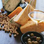 Verschiedene Käsescheiben mit sichtbaren Löchern und blauen Adern, umgeben von Walnüssen, Haselnüssen, einer Schale mit Oliven, einem Weinglas und einer kleinen Schale mit dunkler Marmelade auf einem Holztisch.