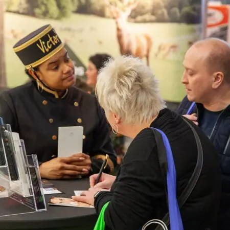 Eine Hotelangestellte in einer Uniform mit der Aufschrift Victoria bedient zwei Kunden an einem Schalter. Der eine Kunde schreibt auf einem Notizblock, während der andere ihm zuhört. Im Hintergrund sind Broschüren und Naturbilder zu sehen.