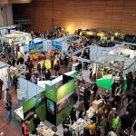 Eine belebte überdachte Kongresshalle mit Menschen, die herumlaufen und verschiedene Ausstellungsstände mit bunten Auslagen und Informationstafeln besuchen. Die Atmosphäre ist lebhaft, und viele Besucher stöbern oder sprechen mit den Ausstellern.