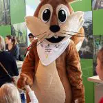 Eine Person in einem großen, freundlichen Eichhörnchenkostüm mit großen Augen und Schnurrhaaren interagiert mit Kindern und Erwachsenen bei einer Indoor-Veranstaltung mit grünen und naturbezogenen Displays im Hintergrund.