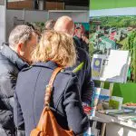 Drei Personen stehen eng beieinander in einem Innenraum und betrachten Broschüren und Hefte auf einem Schautisch. Das Sonnenlicht scheint auf sie, und im Hintergrund ist ein großes Luftbild eines grünen Campus zu sehen.