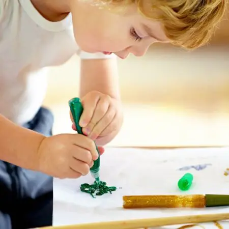 Ein kleines Kind zeichnet mit einem grünen Glitzerklebestift auf ein weißes Blatt Papier und konzentriert sich dabei auf sein Kunstwerk. Ein unverschlossener goldener Glitzerklebestift liegt neben dem Papier.