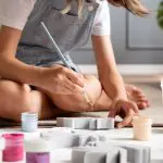 Ein Kind in Jeans-Overall sitzt auf dem Boden, hält einen Pinsel in der Hand und malt geformte Gegenstände. In der Nähe stehen offene Farbdosen in verschiedenen Farben. Die Szene deutet auf eine kreative künstlerische oder handwerkliche Tätigkeit hin.