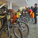 Menschen stehen und begutachten Fahrräder, die in einem Ausstellungsraum ausgestellt sind. Mehrere Fahrräder sind aufgereiht und mit auffälligen SALE!-Schildern versehen. Die Umgebung wirkt geschäftig, mit einer modernen Inneneinrichtung und großen Fenstern.