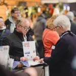 Ein älterer Mann, der eine Broschüre in der Hand hält, spricht mit einer Frau an einem Informationsschalter, umgeben von anderen Menschen in einem belebten Innenbereich. Beide scheinen in ein Gespräch vertieft zu sein, und auf dem Tresen liegt Informationsmaterial aus.