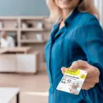 Eine Frau in einem blauen Hemd lächelt und hält ein Veranstaltungsticket mit der Aufschrift "Saarlandhalle" für die Termine 21. und 22. Februar in die Höhe. Der Hintergrund zeigt eine unscharfe Büroumgebung mit Regalen und einer Person, die an einem Schreibtisch sitzt.