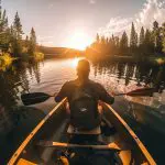 Eine Person mit Rucksack paddelt in einem Kanu auf einem ruhigen See bei Sonnenuntergang, der von Bäumen umgeben ist und das goldene Sonnenlicht auf dem Wasser reflektiert.