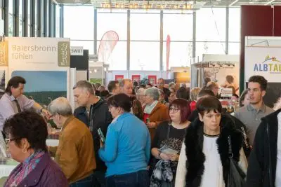 Eine große Gruppe von Menschen geht durch eine belebte Ausstellung oder Messe und sieht sich die Stände mit Informationsschildern und Displays an. Die Atmosphäre ist lebhaft und gut beleuchtet, mit natürlichem Licht, das aus großen Fenstern kommt.