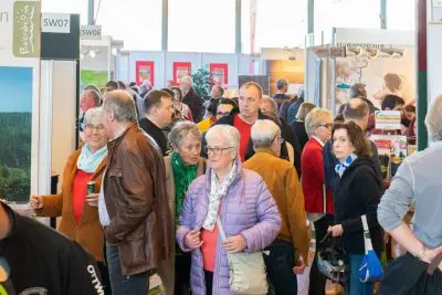 Eine große Gruppe von Menschen, meist ältere Erwachsene, geht durch eine belebte Veranstaltungs- oder Ausstellungshalle, in der im Hintergrund verschiedene Stände und Auslagen zu sehen sind.