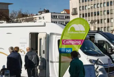 Menschen stehen in der Nähe eines weißen Wohnmobils, das neben städtischen Gebäuden geparkt ist. Auf einer grünen Fahne steht Frankana Freiko Camping ist bunt. Das Wetter ist sonnig und klar.