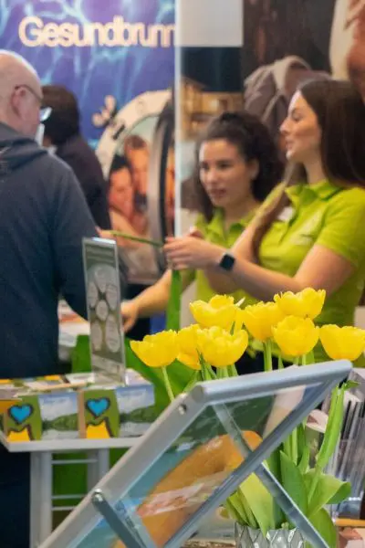 Zwei Frauen in grünen Hemden stehen hinter einem Stand mit gelben Tulpen und Informationsmaterial und unterhalten sich mit einem Besucher einer Indoor-Veranstaltung oder Ausstellung.