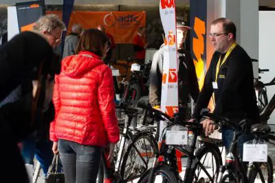 Eine Gruppe von Menschen, darunter ein Mann in einem schwarzen Hemd mit einem gelben Schlüsselband, versammelt sich in einem Innenraum um mehrere Fahrräder auf einer Fahrradausstellung oder -messe, wie es scheint. Im Hintergrund sind Schilder und Transparente zu sehen.
