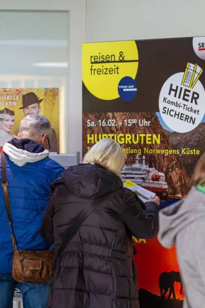 Zwei Personen, die sich den Rücken zuwenden, interagieren mit einem Kiosk oder Fahrkartenautomaten. Ein großes Plakat neben ihnen wirbt für eine Reise- und Freizeitveranstaltung mit einem Kreuzfahrtschiff und fetter gelber und schwarzer Schrift in deutscher Sprache.