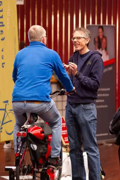 Zwei Männer unterhalten sich bei einer Indoor-Veranstaltung; der eine sitzt auf einem stationären Fahrrad und schaut weg, während der andere vor ihm steht und lächelt und gestikuliert. Im Hintergrund sind Banner und Informationsdisplays zu sehen.