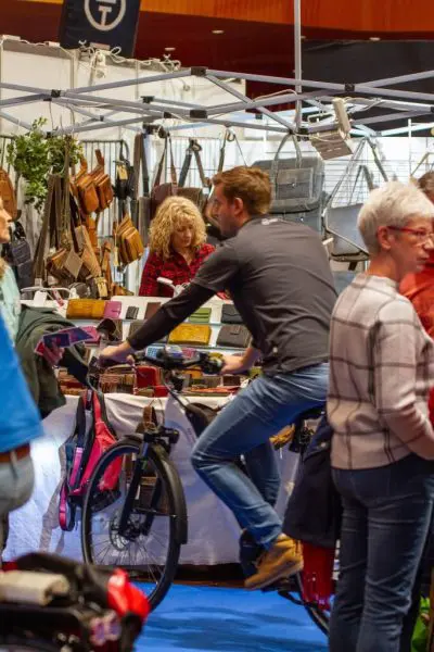 Ein Mann fährt auf einem stationären Fahrrad an einem belebten Marktstand, umgeben von Menschen, die Lederwaren und Accessoires auf Tischen ansehen. Eine Frau steht hinter dem Stand, und die Kunden laufen in der Nähe.