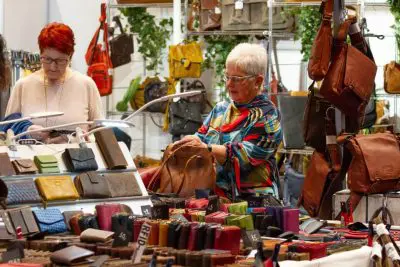 Zwei Frauen stöbern an einem Marktstand in Ledertaschen und Geldbörsen. Eine Frau trägt eine Brille und einen bunten Schal, die andere hat kurzes rotes Haar. Verschiedene Geldbörsen und Handtaschen sind auf Tischen ausgestellt und hängen hinter ihnen.