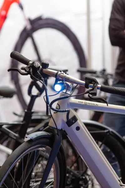 Nahaufnahme eines modernen weißen Fahrrads mit eingeschaltetem Vorderlicht, das den Lenker und einen Teil des Rahmens zeigt. Ein weiteres Fahrradrad ist im unscharfen Hintergrund zu sehen.