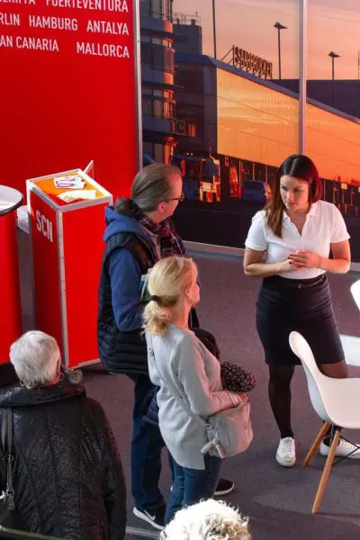 Eine Frau in einem weißen Hemd steht und spricht mit drei Personen in einem Innenraum mit großen Fenstern, die ein Flughafenterminal und Busse draußen zeigen. Ein rotes Schild mit Städtenamen und ein weißer Stuhl stehen in der Nähe.