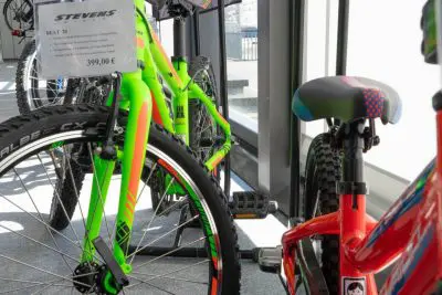 Zwei bunte Fahrräder, ein grünes und ein rotes, sind in einem Innenraum in der Nähe großer Fenster ausgestellt. Auf dem grünen Fahrrad steht ein Schild mit der Aufschrift 399,00 €. Sonnenlicht strömt durch die Fenster und beleuchtet die Fahrräder.