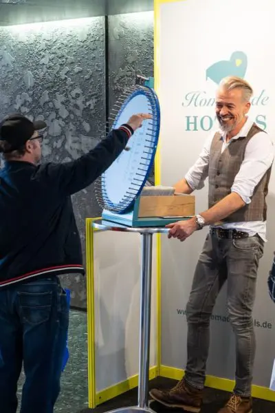 Ein lächelnder Mann steht neben einem sich drehenden Preisrad, während eine andere Person es dreht. Der Mann trägt eine Weste und hochgekrempelte Ärmel, und beide interagieren in einer hell beleuchteten Werbeumgebung.