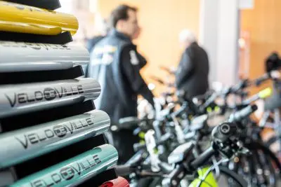 Nahaufnahme von gestapelten VELDEBILLE-Fahrradrahmen in verschiedenen Farben mit unscharfen Menschen und zusammengebauten Fahrrädern im Hintergrund, was auf ein Fahrradgeschäft oder eine Messe hindeutet.