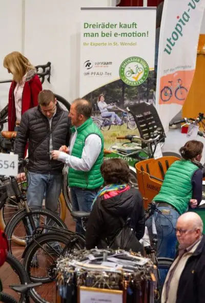 Menschen auf einer Indoor-Fahrradausstellung begutachten Fahrräder. Zwei Männer in grünen Westen unterhalten sich neben Fahrrädern und einem Banner, das für Dreiräder von "e-motion" wirbt. Andere Besucher sehen sich Fahrräder und Displays im Hintergrund an.