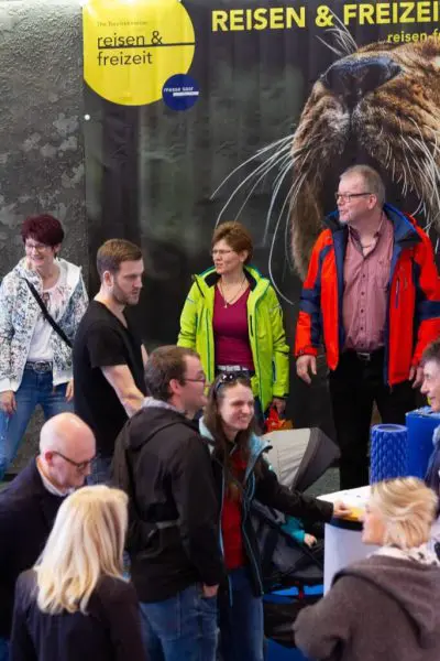 Eine Gruppe von Menschen steht auf einer Reise- und Freizeitveranstaltung und tauscht sich aus. Im Hintergrund ist ein großes Plakat mit einer Nahaufnahme eines Tieres und der Aufschrift Reisen & Freizeit zu sehen.