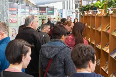 Eine Gruppe von Menschen steht dicht beieinander vor einem hölzernen Regal, das mit Büchern oder Zeitschriften gefüllt ist, auf einer Indoor-Veranstaltung, z. B. einer Messe oder Ausstellung, mit Bannern im Hintergrund.
