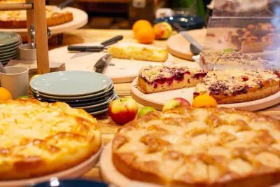 Ein mit verschiedenen Obstkuchen und -torten gefüllter Tisch, umgeben von frischen Äpfeln, Orangen, Stapeln von Tellern und Servierutensilien. Die Desserts sind teilweise aufgeschnitten und bereit zum Servieren.