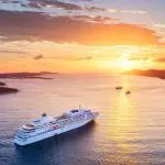 Ein großes weißes Kreuzfahrtschiff fährt bei Sonnenuntergang auf ruhigem Wasser, mit einem leuchtend orangefarbenen Himmel, vereinzelten Wolken und entfernten Inseln am Horizont. Zwei kleinere Schiffe sind im Hintergrund zu sehen.