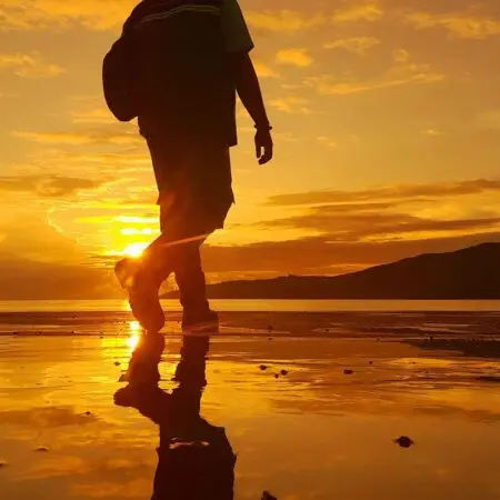 Silhouette einer Person, die bei Sonnenuntergang am Strand spazieren geht, wobei sich das goldene Sonnenlicht auf dem nassen Sand und dem ruhigen Wasser spiegelt und die Hügel im Hintergrund sichtbar sind.