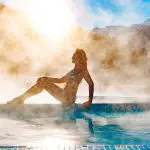 Eine Frau im Bikini sitzt am Rande eines Freibads, umgeben von Dampf, mit Bergen und hellem Sonnenlicht im Hintergrund.