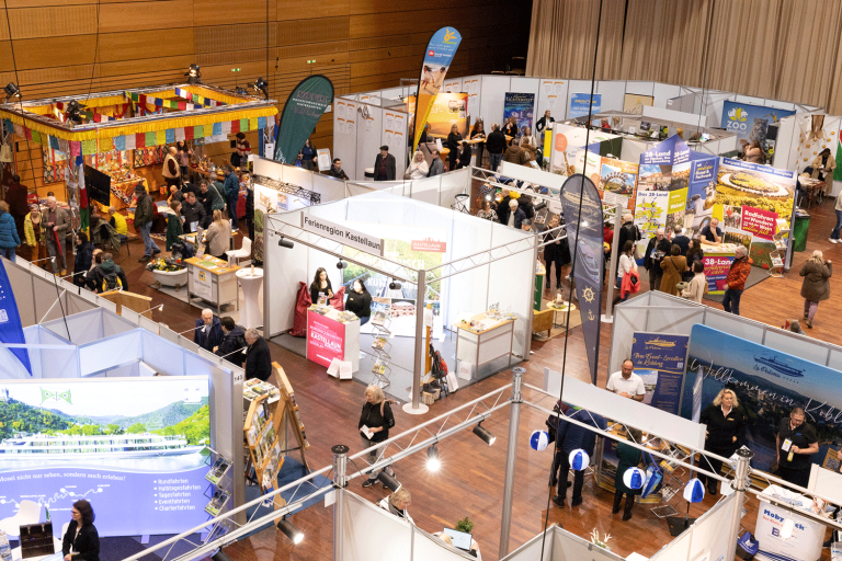 Eine belebte überdachte Ausstellungshalle mit zahlreichen Ständen, Bannern und Displays. Die Leute gehen umher, unterhalten sich und schauen sich die verschiedenen Stände an. Die Szene ist lebendig, mit bunten Dekorationen und verschiedenen Werbematerialien.