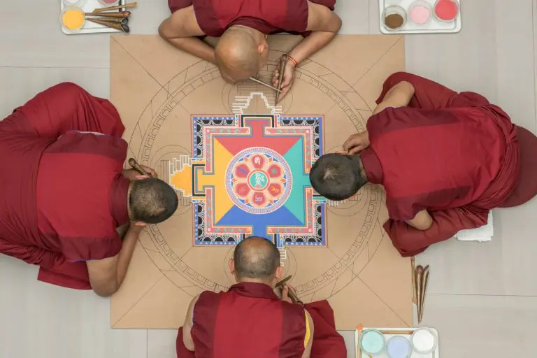 Vier Mönche in roten Roben erstellen ein farbenfrohes Sandmandala auf einer hellbraunen Fläche. Sie arbeiten mit feinen Werkzeugen und Farbschalen, von oben gesehen.