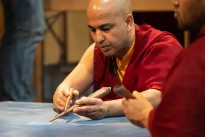 Ein Mann in rotem Gewand arbeitet konzentriert mit traditionellen Werkzeugen an einem detaillierten Kunstwerk auf einer blauen Fläche, wobei eine weitere Person in rotem Gewand teilweise neben ihm zu sehen ist.