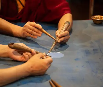 Drei Hände arbeiten mit traditionellen Metallwerkzeugen vorsichtig ein Sandmandala auf einer blauen Oberfläche, wobei sie sich auf einen kleinen Bereich mit blauem Sand konzentrieren. Im Hintergrund befindet sich eine Messingschale.
