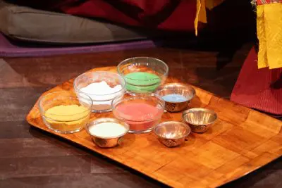Auf einem Holztablett stehen kleine Glasschalen mit bunten Pulvern - gelb, weiß, grün und rot - neben Metallschalen auf einem dunklen Boden, auf dem im Hintergrund Stoff zu sehen ist.