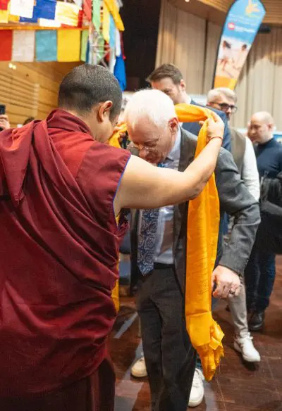 Ein buddhistischer Mönch legt einem älteren Mann im Anzug bei einer Indoor-Veranstaltung einen gelben Zeremonienschal um den Hals, während im Hintergrund Menschen zusehen und bunte Banner wehen.
