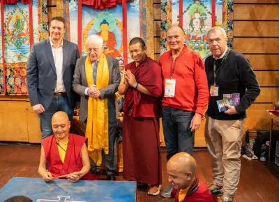 Sechs Männer posieren gemeinsam in einem Innenraum vor einem farbenfrohen tibetisch-buddhistischen Hintergrund. Zwei Mönche sitzen vorne und arbeiten an einem Sandmandala, vier Männer stehen lächelnd hinter ihnen.