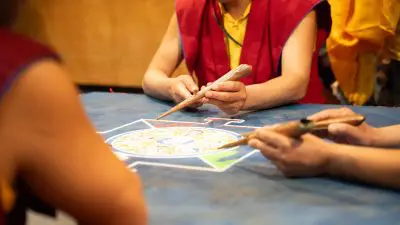 Drei Personen in roten und gelben Gewändern arbeiten mit traditionellen Werkzeugen an einem farbenfrohen Sandmandala auf einer blauen Fläche und konzentrieren sich dabei ganz auf ihr kompliziertes Design.