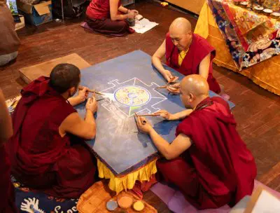 Drei buddhistische Mönche in roten Roben sitzen um einen Tisch und formen mit Metallwerkzeugen sorgfältig ein buntes Sandmandala. In der Nähe sind auf einem Holzboden Kunstgegenstände und bunte Stoffe zu sehen.
