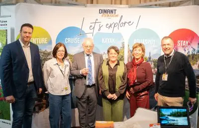 Sechs Personen stehen zusammen und lächeln vor einem farbenfrohen Hintergrund, auf dem Dinant Evasion Lets explore! steht und auf Aktivitäten wie Abenteuer, Kreuzfahrt, Kajak, Wassersport und Aufenthalt hingewiesen wird. Ein Tisch mit Broschüren steht vor ihnen.
