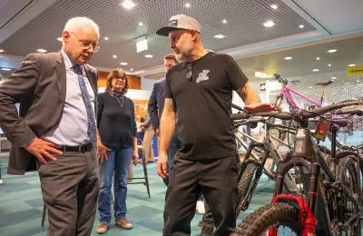 Ein Mann im Anzug hört einem anderen Mann in Freizeitkleidung zu, der bei einer Indoor-Veranstaltung die Funktionen eines Fahrrads erklärt. Im Hintergrund sind zwei weitere Personen und mehrere Fahrräder zu sehen.
