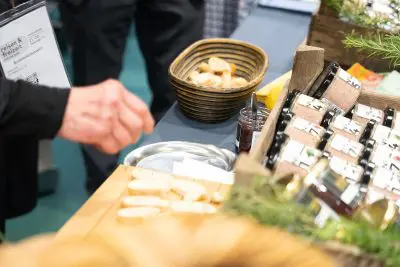 Eine Person greift nach Brotscheiben auf einem Holzbrett an einem Verkostungstisch, auf dem auch Pastete, ein Glas Marmelade und ein Korb mit weiteren Brotscheiben stehen.
