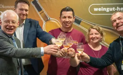 Fünf lächelnde Personen stoßen mit Weißweingläsern vor einem farbenfrohen Hintergrund mit dem Schriftzug "weingut-he" und dem Bild einer Flasche an. Vier Personen sind leger gekleidet, eine trägt einen Anzug.