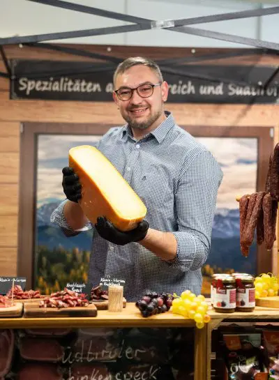 Ein lächelnder Mann mit Brille und schwarzen Handschuhen hält einen großen Käseblock an einem Marktstand mit Fleisch, Weintrauben und Gläsern. Hinter ihm sind ein Schild in deutscher Sprache und eine malerische Bergkulisse zu sehen.